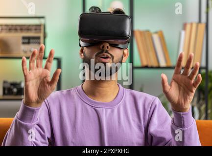 Indian man using virtual reality futuristic technology VR app headset helmet to play simulation 3D 360 video game, watching film movie at modern home apartment. Hindu guy in goggles sitting on couch Stock Photo