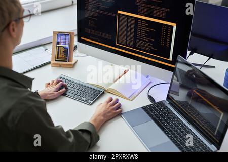 Rear view of young programmer writing security codes on computer connecting laptop and smartphone sitting at his workplace Stock Photo