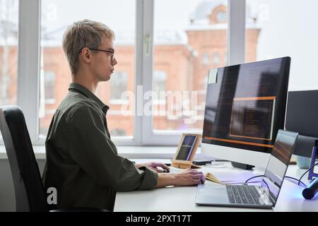 Side view of young programmer sitting at his workplace and working with security codes for program using computer and smartphone Stock Photo