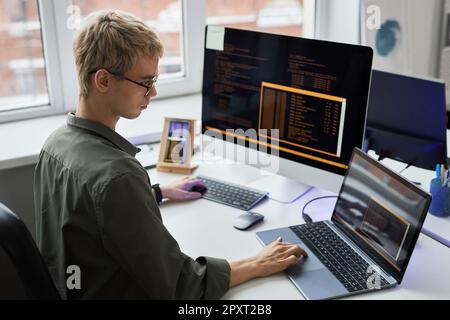 Rear view of young programmer in eyeglasses writing codes on different gadgets while working at his workplace in IT office Stock Photo