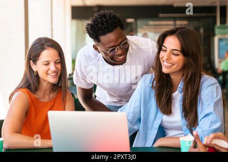 Group of happy multiracial students sitting in a cafe bar looking at laptop. Three cheerful friends studying together with a computer Stock Photo