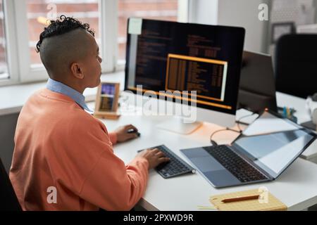 Rear view of young developer writing security codes on computer connecting it with laptop and smartphone while sitting at his workplace in office Stock Photo