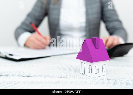 Businesswoman sitting and writing in notebook. Paper house' on desk. Stock Photo