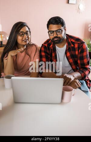 Happy indian family couple cuddle at desk make video call to friends using laptop webcam. Loving young spouses look at computer screen waving hands in Stock Photo
