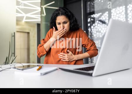 Businesswoman has severe nausea, latin american woman at work sick, working sitting at table with laptop inside office Stock Photo