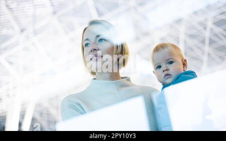 Thoughtful young mother looking trough window holding his infant baby boy child while waiting to board an airplane at airport terminal departure gates Stock Photo