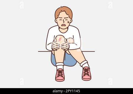Crying teenager boy sits on ground and holds on to injury on knee after fall during walk. Unfortunate child was injured during sports activities or ac Stock Photo
