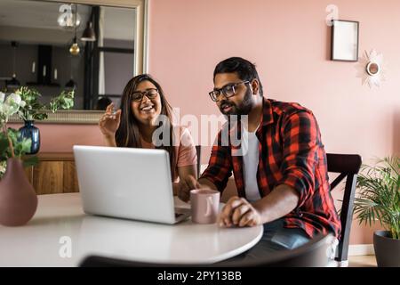 Happy indian family couple cuddle at desk make video call to friends using laptop webcam. Loving young spouses look at computer screen waving hands in Stock Photo