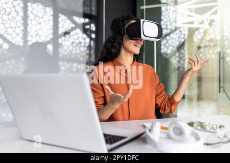 Successful latin american woman in office using vr glasses for video call and chatting online in virtual reality simulator, online conference with colleagues remote in metaverse. Stock Photo