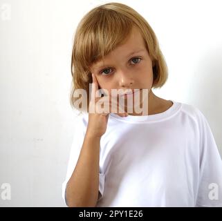 Little cute girl 6-7 years old with blond hair on a white background. Tanned skin. White T-shirt. Copy space. The girl raised her right hand and put h Stock Photo