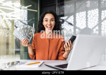 Online casino. Young muslim woman sitting in office at table with laptop, holding cash in hand and phone, playing online games, making bets. Smiling at the camera, celebrating the win. Stock Photo