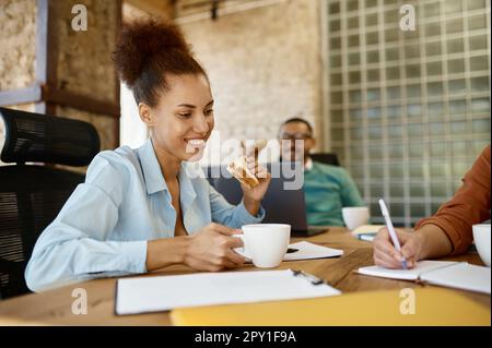 Multiracial marketing team having meeting during lunch time in coworking office. Focus on young woman eating sandwich and drinking coffee Stock Photo