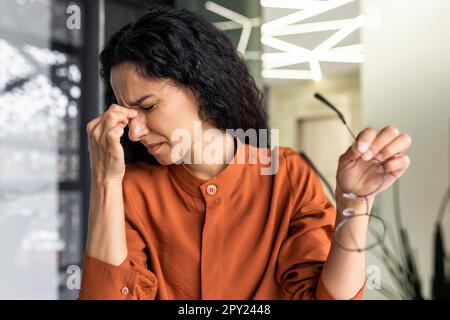 Overworked hispanic business woman working inside modern office building, female worker removed glasses and rubs eyes, uses laptop. Stock Photo