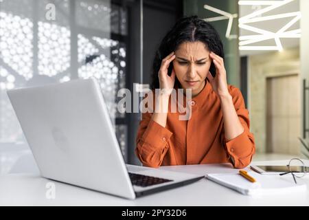 Overtired hispanic business woman working inside modern office, office worker with laptop having severe headache Stock Photo