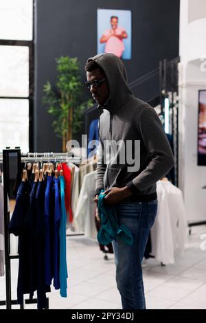 African american man thief looking at trendy clothes before stealing it in modern boutique. Robber wearing sunglasses and hood not be recognized in clothing store. Burglary concept Stock Photo