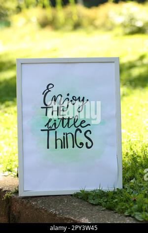Poster with phrase Enjoy The Little Things on grass outdoors Stock Photo