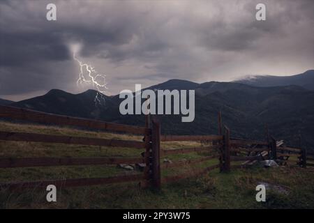 Dark cloudy sky with lightnings over mountain countryside with wooden fence. Thunderstorm Stock Photo