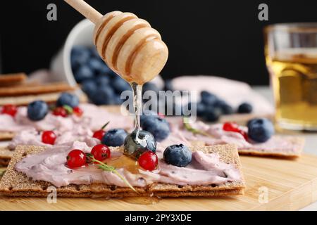 Pouring honey onto tasty cracker sandwich with cream cheese, blueberries, red currants and thyme on wooden board, closeup Stock Photo