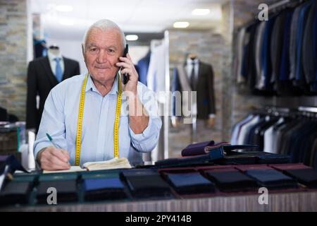 Professional tailor communicates with the client on mobile phone and takes notes in notebook Stock Photo