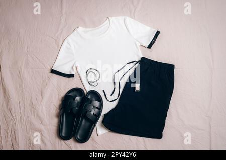 White T-shirt, black shorts, black slippers, necklace and nail polish on grey background. Overhead view of woman's casual day outfit. Trendy stylish s Stock Photo