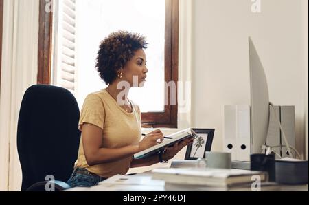 Working tirelessly to reach success. a young businesswoman working on a computer while going through notes from a book in an office. Stock Photo