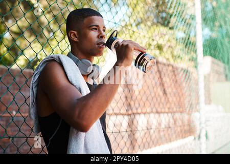 Taking a breather to get some energy. a sporty young man drinking water while standing against a fence outdoors. Stock Photo