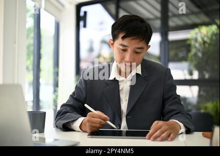 Focused millennial Asian businessman planning his new project on tablet at his desk. Stock Photo