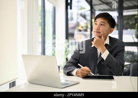 Smart and inspired millennial Asian businessman thinking, pondering and planning his business project at his desk. Stock Photo
