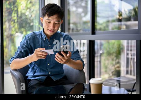 Happy and handsome millennial Asian man using mobile banking application to pay his bills online. Stock Photo