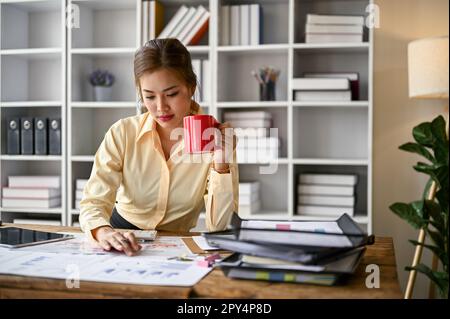 Focused and professional young Asian female accountant or financial worker sipping coffee and analyzing business financial data on reports at her desk Stock Photo