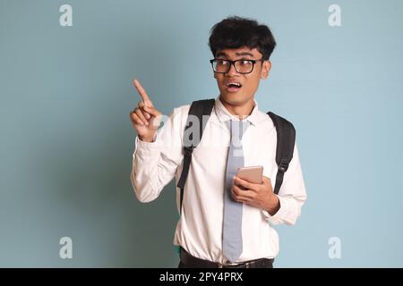 Indonesian senior high school student wearing white shirt uniform with gray tie showing and presenting mobile phone while pointing away with finger. I Stock Photo