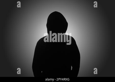 Mysterious man wearing black hoodie standing against dark background. Hacker, crime, and cyber security concept. Stock Photo