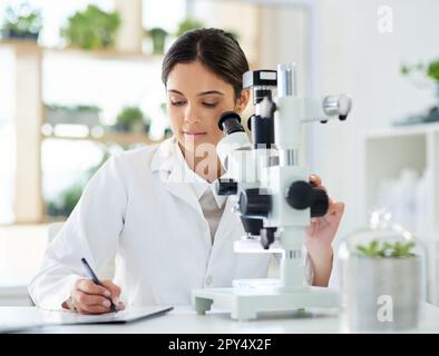 Noting down a new breakthrough. a young scientist writing notes while using a microscope in a lab. Stock Photo