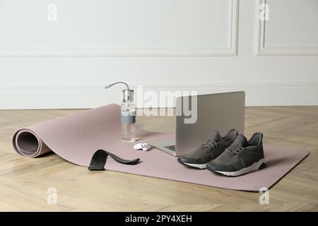 Exercise mat, laptop, bottle of water, wireless earphones, fitness elastic band and shoes on wooden floor indoors Stock Photo