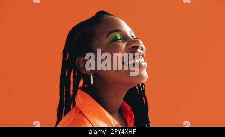 African woman with makeup and face piercings smiling with her eyes closed. Black woman standing against an orange background. Stock Photo