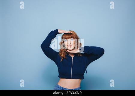 Playful young woman framing her face with her hands in a studio. Woman with ginger hair having fun while standing against a blue background. Stock Photo