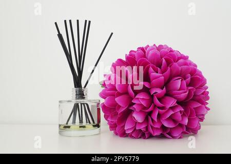 Arrangement with incense sticks, essential oil in a glass vase and a bright pink-lilac terry artificial flower in the interior of a white room. White background Stock Photo