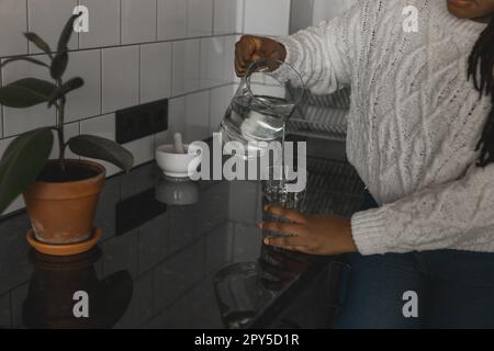Close-up thirsty african american woman drinking mineral water in kitchen, pouring healthy liquid from jug to glass, happy lady with braids enjoying refreshing drink Stock Photo