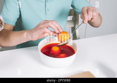 Cropped photo of boy hands, coloring orange Easter hen eggs in spoon, bowl, table. Soak up, absorption color. Close up Stock Photo
