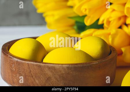 Easter eggs in wooden bowl and bouquet of bright yellow tulips on white wooden surface on gray background Stock Photo