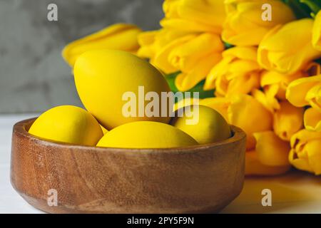 Easter eggs in wooden bowl and bouquet of bright yellow tulips on white wooden surface on gray background Stock Photo