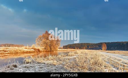 Dawn on winter riverbank. Frost on grass, cane. Snowy rural road. January forest river sunrise. Cold Weather landscape, reflection in water. Stock Photo