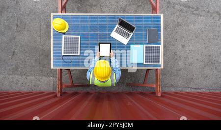 Shipment worker in hardhat and safety vest looking at white screen tablet computer monitor. Planning  the delivery schedule for the next shipment. He use solar cell panel as a table. Top View Stock Photo