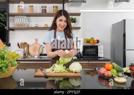 Young asian housewife dressed in an apron peeling fresh lettuce on wooden chop board. The kitchen counter full of various kinds of vegetables. Stock Photo
