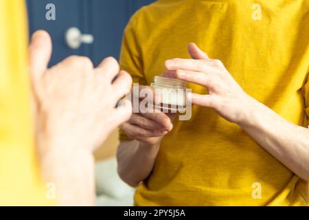 Hands of middle aged man holding jar of moisturizing cream. Crop male portrait in room mirror. Skincare morning routine. Stock Photo