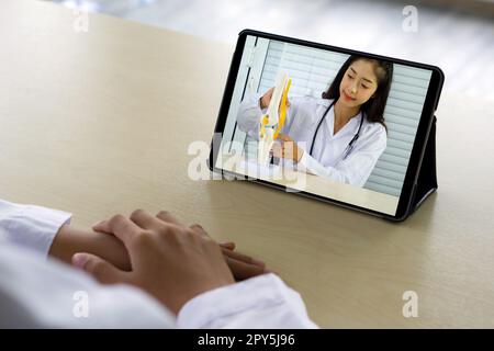Back view of woman in white gown have webcam conference on tablet computer with young asian doctor  about bone, joint, tendon and muscle. Stock Photo