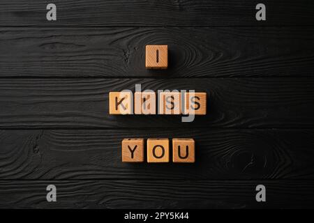 Wooden cubes with the phrase I Kiss You on a black background - a wooden table. Words of kiss are made of letters from wood for your girlfriend, boyfriend, wife, husband, for Valentine's Day greeting romantic postcard. Stock Photo