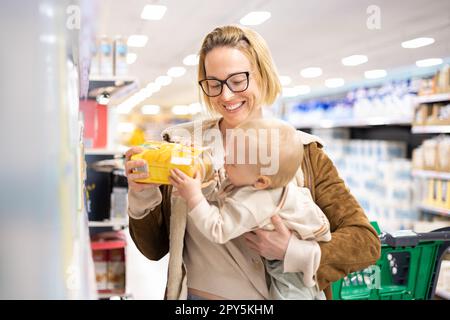 Caucasian mother shopping with her infant baby boy child choosing products in department of supermarket grocery store. Stock Photo