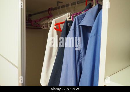 Men's shirts hang on hangers in an open white closet. Men's fashion. Organization of things in a closet or dressing room. Blue and white shirts. Housekeeping. Stock Photo