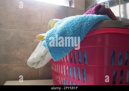 Towels in the laundry basket. Blue and pink cotton terry towels are thrown into a pink plastic basket. Housekeeping. Storing and separating laundry before washing. Light from above from open window. Stock Photo